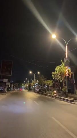 The video shows a street at night, illuminated by streetlights. The road is wet, suggesting it has recently rained. There are trees lining the right side of the road, and buildings on both sides. A few vehicles can be seen in the distance. The camera appears to be moving, possibly from a vehicle. The overall atmosphere is quiet and deserted.