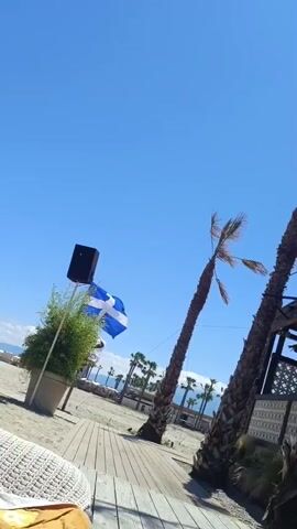 A wide shot of a beach scene on a bright sunny day. In the foreground, a white woven lounge chair with a yellow and red towel draped over it sits on a wooden deck. To the left of the chair, a large potted plant with green foliage stands next to a black speaker. A Greek flag waves gently in the background. Palm trees line the sandy beach, and in the distance, beach chairs and umbrellas are visible, along with some buildings and a clear blue sky. The camera is angled upwards, capturing the expanse of the sky.