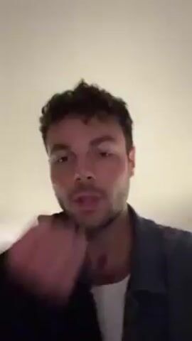 The video shows a man named Tyler, with curly dark hair and a denim jacket, who is speaking directly to the camera. He identifies himself and greets the "collective memory gang". In the background, another man with a beard is visible in what appears to be a kitchen setting. The lighting is dim, and the ceiling has a recessed light fixture. Tyler is gesturing with his hand as he speaks.