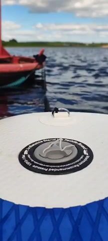 The video is a close-up, low-angle shot of a white paddleboard with a blue trim. In the foreground, a gray air valve is visible on the paddleboard, and further back, a metal D-ring is attached to the board. Beyond the paddleboard, the water is calm and dark blue. A red buoy floats in the distance, tethered by a rope. The sky is partly cloudy with a bright blue hue. The focus is on the paddleboard and its valve, with the buoy and water in the background.