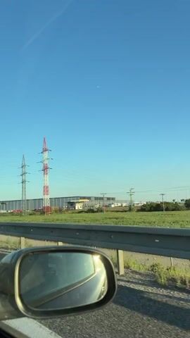 The video is a first-person perspective from a moving vehicle, likely a car, looking out at a landscape. The shot begins with a clear blue sky and two large metal power line towers in the distance. One tower is red and white, the other is silver. A large, modern warehouse-like building is situated behind the towers. The building has "BILLA" and "REWE" logos visible on its side, suggesting it is a retail or logistics center. The foreground of the shot shows a car's side-view mirror, reflecting the sky and a bit of the vehicle's interior. A highway guardrail runs along the bottom edge of the frame. In the sky, a small red object, possibly a balloon or a drone, is visible. The video appears to be filmed during daylight hours, with bright sunlight. The movement of the vehicle suggests travel along a road.