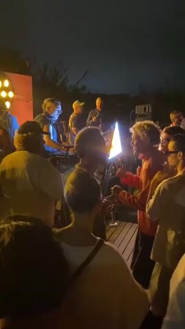The video captures a lively outdoor gathering at night. A group of people are clustered together, with some individuals on a raised platform or stage area. Several people are holding cups, and one person is holding a yellow cup aloft. One person on the stage appears to be speaking into a microphone, while others are interacting with musical equipment, suggesting a performance or DJ set. The scene is illuminated by stage lighting, including a prominent spotlight and a circular light fixture. The atmosphere appears festive and energetic, with people gathered closely and engaged in the event. The background is dark, indicating it is nighttime, with some faint greenery visible.