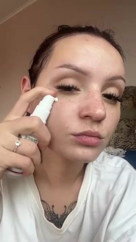 A young woman with dark hair pulled back is applying white cream to her face from a white pump bottle. She is wearing a white t-shirt. She has freckles and long eyelashes. The background shows a dimly lit room with a bed and curtains.