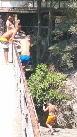 A group of people stand on a bridge looking down at a narrow body of water. Some are wearing swim trunks, and one person is preparing to jump into the water. The bridge appears to be made of metal and concrete. The surrounding area is covered in trees and rocks. The water is murky.