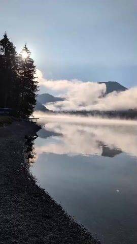 The video displays a serene lakeside landscape during sunrise. The sun casts a warm glow on the scene, illuminating a line of trees on the left side of the frame. The lake's surface is calm, reflecting the misty mountains and clouds in the background. A thin layer of mist hovers above the water, creating an ethereal atmosphere. The foreground features a pebbled shoreline. The camera pans slowly to the right, revealing more of the lake and the surrounding mountains.