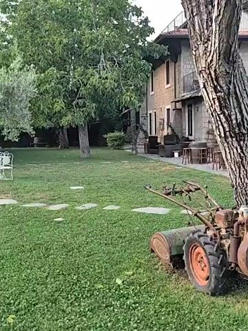 This is a video of a rural landscape with a building and a grassy yard. The building appears to be a house or a restaurant, with a stone facade and a wooden roof. There are trees, an olive tree, and a table with chairs in the yard. A tractor is visible in the background. The scene is set in a hilly area, with a forest in the background. The time of day appears to be late afternoon or early evening, with a cloudy sky. The video is shot from a fixed position, with no movement.