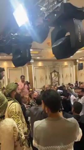 The video shows a large group of people gathered in what appears to be a ballroom or large reception hall, with stage lights illuminating the scene. Many individuals are wearing traditional Middle Eastern attire, including headscarves and robes. Some people are clapping and dancing, suggesting a celebration or event. Numerous camera operators and photographers are present, recording the occasion. The environment is ornate, with decorative wall panels and detailed ceiling work. The focus seems to be on a specific group of people in the center, possibly the guests of honor.