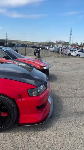 The video shows a bright red Toyota Chaser with a black carbon fiber hood and body kit, parked in a gravel parking lot. The car has sponsor logos on the side and aftermarket wheels. The camera pans along the side of the car, showing the interior roll cage and other cars parked in the background. People are walking around in the parking lot.