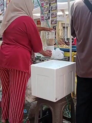 A woman wearing a red shirt and red and white striped pants, and a light brown hijab, stands behind a white blender. She is holding the blender's lid and operating the machine, which appears to be blending a light brown liquid. In front of the blender, a white styrofoam cooler sits on a brown plastic stool. The woman is likely at an outdoor market, as indicated by the stalls and items for sale visible in the background. A person's back is visible on the right side of the frame.
