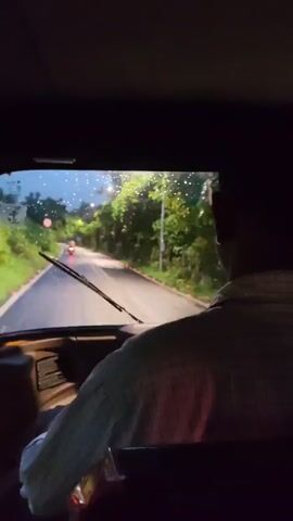 The video shows the view from inside a vehicle looking out the windshield at a wet road at dusk or night. Raindrops are visible on the windshield and on the road. The headlights of the vehicle illuminate the road ahead, where a person on a scooter is riding away from the camera. Lush green trees line both sides of the road, and there are streetlights visible in the distance. A speed limit sign is partially visible on the left side of the road. The driver's shoulder and the back of their head are visible in the foreground on the right.