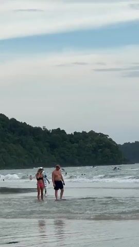 Two adults, a man and a woman, are standing in shallow ocean water on a beach, with a forested hill in the background. The man is wearing dark shorts and is facing towards the right. The woman is wearing a pink and black bikini and is facing towards the left. Further out in the water, several jet skis are being ridden by people. The sky is overcast with some blue patches visible. The waves are gentle.