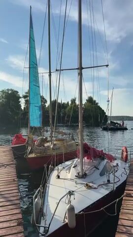 The video shows a view from a wooden dock looking out at several sailboats on a body of water. In the foreground, two sailboats are docked side-by-side, with a wooden plank walkway extending from the dock to the second sailboat. The sailboat on the left has a large, bright blue sail with white markings, while the sailboat on the right is mostly white with a red hull and a covered sail. Both sailboats have tall masts and rigging. In the background, more sailboats are visible on the water, along with a small boat with people on it and a distant treeline. The sky is blue with scattered white clouds, and the water is calm with gentle ripples. The scene appears to be set in the late afternoon or early evening, with the sun casting a warm glow. The overall atmosphere is peaceful and serene, suggesting a leisurely day on the water.