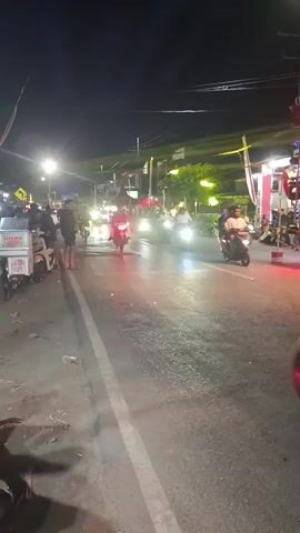 A nighttime street scene shows multiple motorcycles and scooters on a road. A few pedestrians are standing on the side of the road near parked motorcycles. The street is illuminated by streetlights and the headlights of the vehicles. There are some buildings and trees visible in the background. The overall atmosphere appears to be busy and lively.