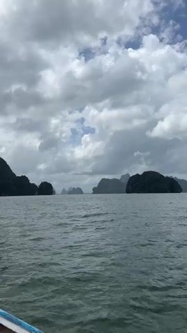 A video shot from a boat shows a wide expanse of choppy gray water with several large, dark, tree-covered islands in the background. The sky above is overcast with heavy gray clouds, but there are patches of lighter sky with some blue peeking through in the upper center of the frame. The islands are rugged and mountainous, with some appearing as sharp peaks and others as rounded, hulking masses. The overall mood of the scene is somber and dramatic due to the stormy sky.