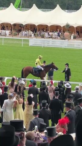 The media file captures a scene at a horse racing event, likely at Ascot, United Kingdom. The foreground is dominated by a crowd of spectators, many dressed in formal attire, including top hats and elaborate hats, suggesting a significant social occasion. The spectators are gathered behind a white railing, looking towards a grassy racecourse.

On the racecourse, a jockey in a bright yellow and black polka-dot racing silk rides a brown horse. The horse is led by a person in a dark suit. In the background, a large, white, tent-like structure with multiple peaks provides shade and houses seating areas where other attendees are visible. This structure, along with the manicured green grass of the racecourse and the trees in the distance, suggests a well-established and possibly prestigious venue.

The time of day appears to be daytime, with overcast or diffused lighting, indicating it might be a cloudy day, or perhaps late afternoon. There is no visible text in the media file. The overall atmosphere seems to be one of anticipation and observation, typical of a sporting event where spectators are keenly watching the proceedings on the track. The presence of formal wear and the grand setting imply a high-society event.