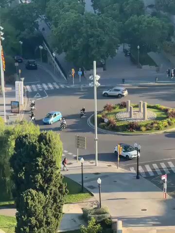 The video presents a high-angle, panoramic view of a coastal city scene, likely Tarragona, Spain, on a clear, sunny day. The lighting suggests it's either morning or late afternoon, with clear shadows cast.

The initial shot focuses on a busy urban intersection featuring a prominent roundabout. Vehicles, including a small blue car, a silver SUV, a white car, and several motorcycles, are seen actively moving through and around the roundabout. A few pedestrians are visible on the sidewalks surrounding the intersection. The area is characterized by green spaces, bushes, and various trees, including prominent palm trees. Street lamps, traffic lights, and poles flying flags with red and yellow stripes (consistent with Catalan or Spanish flags) are also part of the urban infrastructure.

As the camera slowly pans upwards and to the right, the perspective widens to reveal more of the coastal environment. A lush green landscape with numerous trees and palm trees stretches along what appears to be a road leading down towards the sea. The final part of the video captures a vast expanse of calm, blue sea under a clear sky, with several large cargo ships or tankers anchored in the distance.