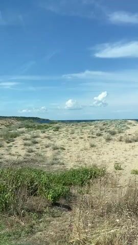 The video shows a car driving through a sandy, natural landscape. The car is silver and is seen from the rear. The landscape consists of sand dunes covered with sparse vegetation. The sky is blue with some white clouds. The overall impression is that of a coastal or desert environment on a sunny day. There are no people visible in the video.