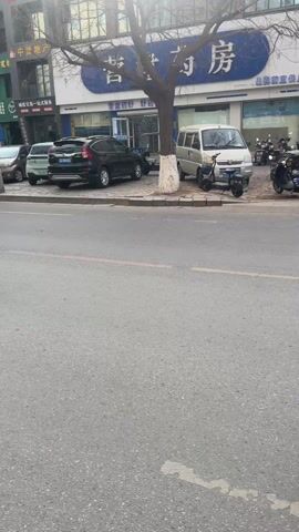 The video shows a street scene in China. There is a blue bus labeled "11路" driving down the road. Several people are standing on the sidewalk watching traffic, including a group of people who are near a metal railing. Many people are riding motorbikes, and there are several cars. The sky is overcast. Some of the storefront signs are written in Chinese.
