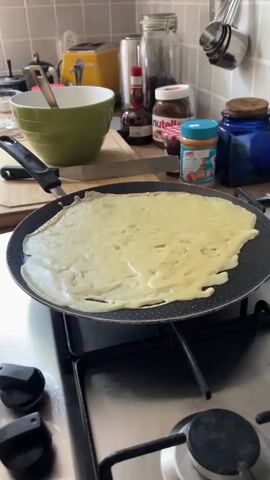 The video shows a close-up of a crepe being cooked in a pan on a stovetop. The crepe is thin and appears to be just starting to set on one side, with uncooked batter visible on the other. The pan is dark-colored and non-stick, with a slight sheen of oil. In the background, out of focus, there are kitchen counter items including jars of Nutella and peanut butter, a green bowl with a whisk, and a toaster. The overall impression is of a home kitchen environment with food preparation in progress.