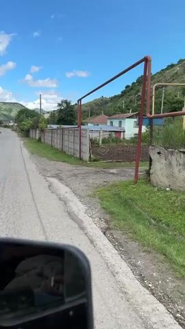 The video shows a view from inside a vehicle traveling down a paved road. The road is cracked and appears to be in a rural area. On the right side of the road, there is a long, grey concrete fence with red metal poles interspersed. Behind the fence, there are houses and trees. On the left side of the road, there is a grassy shoulder and more trees and houses further in the distance. The sky is blue with white clouds. In the foreground, a car's side mirror is visible, reflecting the interior of the vehicle. The video is shot from a moving vehicle, indicated by the slight blur and the changing perspective.