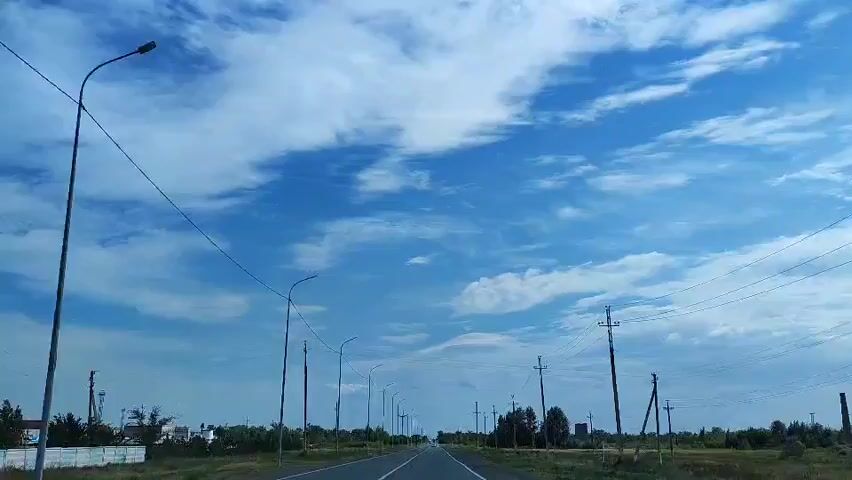 This video shows a forward-facing view from a vehicle moving along a two-lane road under a bright blue sky with scattered clouds. The road is paved with asphalt and has a white dashed line dividing the lanes. On either side of the road, there is dry grass and some trees. Several utility poles with wires run alongside the road, and there are also streetlights visible. In the distance, a few buildings can be seen to the left, possibly industrial structures. The overall impression is a drive through a rural or semi-rural landscape on a clear day.
