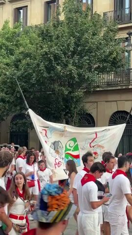 The video shows a group of people, mostly young adults, walking outdoors in what appears to be a festival or parade. They are all dressed in white shirts and shorts, with many wearing red sashes or neckerchiefs. Two large white banners with colorful illustrations are being held aloft by poles, carried by individuals within the crowd. The banners depict various figures, including what looks like a bull and other stylized characters, along with abstract shapes and text. The setting is an urban environment with buildings in the background, one of which has arched windows. A large, leafy tree is visible in the upper left portion of the frame. The people are interacting with each other, some talking, others looking at their phones, and some are holding drinks. The overall atmosphere seems festive and lively.