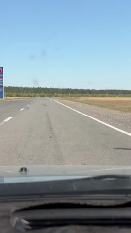 The video shows a view from inside a car looking forward at a paved road. The road is empty and appears to be in a rural or semi-rural area, with trees and fields visible on either side. The sky is clear and blue. The car is in motion, indicated by the slight blur of the scenery. The dashboard of the car is partially visible at the bottom of the frame.