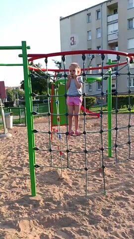 A young girl with blonde hair, wearing a striped t-shirt and pink shorts, is climbing a rope net structure on a sandy playground. She is holding onto the ropes with her hands and her feet are placed on the net. The playground equipment is green and red. In the background, there is a building with the number "3C" visible on it. The sky is clear and bright, suggesting it is daytime. The overall scene appears to be a sunny day at a playground, likely in Kolno, Poland, given the context. The girl seems to be actively engaged in climbing, with no clear indication of her emotional state beyond focused activity. The setting is an outdoor recreational area.
