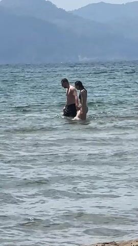 The video depicts a man and a woman standing in the sea. The man is fair-skinned, wearing dark swimming trunks and is shirtless. The woman has dark hair and is wearing a two-piece dark swimsuit. They are standing in the water up to their thighs. In the background, there's a mountain range under a clear sky. The water is light turquoise and slightly rippled.