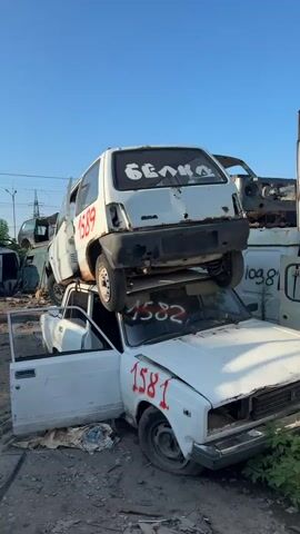 The video shows a junkyard with many old cars. In the foreground, there are two white cars stacked on top of each other. The bottom car has its driver's side door open, and red numbers are spray-painted on both cars. The top car has the word "БЕЛКА" (which translates to "Squirrel" in Russian) spray-painted on its rear window. In the background, there are large piles of mangled car parts and more stacked vehicles. The sky is clear and blue, and the sun appears to be shining. The ground is covered in dirt and debris. There are no people visible in the video.