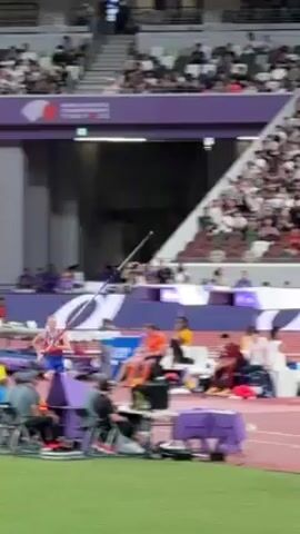 At an athletic stadium with a packed audience, a man is pole vaulting. He wears red, white, and blue athletic attire. He runs and plants the pole, then vaults himself over the bar, landing on a cushioned mat. Many people are in the background and various items like cameras and bags are sitting near the edge of the athletic field.