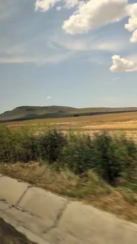 A stationary camera captures a landscape scene from a moving vehicle. The foreground shows a concrete curb and asphalt. Beyond that, a stretch of dry grass and shrubs is visible, with a distinct concrete barrier running along the edge of the road. In the mid-ground, a large, flat, harvested field of dry, golden crops stretches out. Further in the distance, a low, rounded hill with a dark, possibly wooded, summit is silhouetted against the sky. The sky is a clear blue with scattered white, puffy cumulus clouds. The overall impression is of an agricultural or rural setting under a bright, sunny sky.