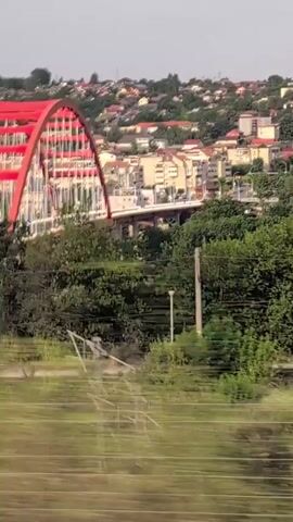 The video shows a view from a moving vehicle, likely a train, looking out at a landscape. In the foreground, there are blurred green trees and some horizontal lines, suggesting a fence or railing. In the midground, there is a town with buildings, trees, and a distinctive red bridge-like structure. The sky is visible in the background. The camera is panning or moving, causing the motion blur. The scene appears to be a daytime view of a semi-urban or suburban area with greenery.