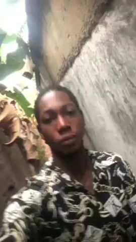 A young man is holding a camera angled upwards at himself while walking through a narrow stone passage with vegetation at the top opening. He is wearing a patterned black and white button-down shirt. The passage walls are grey and weathered. Natural light filters in from the opening above. The man appears to be the only person in the video. He looks directly at the camera as he moves forward through the passage.