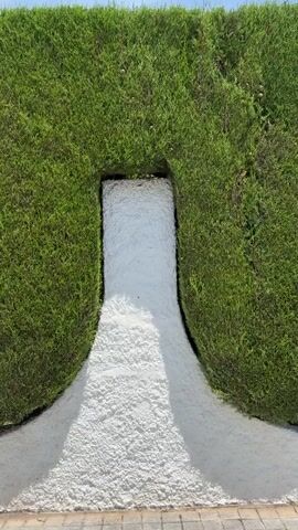 A still image of a water feature, with water flowing over a light-colored, textured surface that is carved into a green hedge. The water flows from a darker, recessed area within the hedge and cascades down, fanning out towards the bottom. The hedge is green and lush, forming a symmetrical opening for the water to pass through.