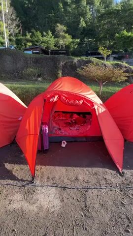 A child is sleeping in a red tent. The child is covered with blankets and is lying on a bed with floral patterned sheets. There is a pink suitcase and a white sandal outside the tent. In the background, there are trees and a hill. The scene appears to be a campsite.