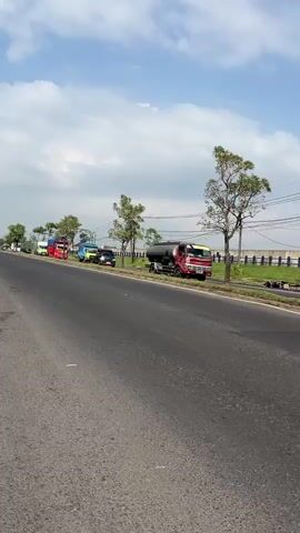 This is a vertical video taken from the side of a road. The road is asphalt and appears to be somewhat worn. On the right side of the road, there is a line of parked trucks. The trucks vary in color and type, including what appears to be a tanker truck and several larger freight trucks. Some of the trucks are brightly colored, red, blue, and green. There are trees and power lines visible beyond the trucks. The sky is blue with scattered white clouds. The scene suggests a roadside area, possibly a parking area for trucks or a place where they are stopped. There are no people clearly visible in the video.