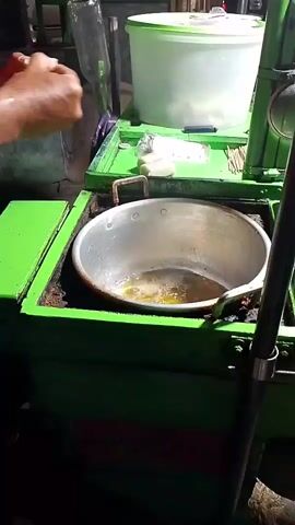 A person's hands are visible in the foreground, preparing food. The person is holding a small white object and appears to be dropping it into a metal pan filled with hot oil. The pan is on a green-colored cooking cart. The background shows other cooking utensils and containers, as well as some lighting that suggests an outdoor or dimly lit environment. The action is focused on the preparation of food, with the hands and the cooking process being the primary subjects.