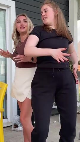 The video shows two young women dancing outdoors. The woman on the left has blonde hair and is wearing a brown top and a white skirt. She is holding a small black object in her right hand. The woman on the right has reddish-blonde hair and is wearing a black t-shirt and black sweatpants. They are both moving their arms and bodies in a rhythmic way. The background appears to be a house with siding and a window. There is also a yellow chair and an air conditioning unit visible. The lighting suggests it is daytime.