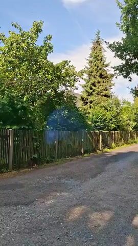 A dirt road is flanked by a wooden fence on the left and trees on the right. Smoke is visible above the fence. The road appears to be in a rural or suburban setting. A tall evergreen tree is visible behind the fence. The sky is blue with some clouds. A red and white circular sign with the number '2' is visible on a pole near the fence. Houses and more trees are visible in the background. The camera pans to the right, showing more of the road, fence, and houses.