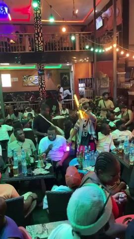 The video captures a lively nighttime scene inside a crowded bar or lounge in Obosi, Nigeria. Numerous people, predominantly adults, are present, socializing at various tables and standing areas across multiple levels of the establishment. The atmosphere is energetic, with artificial lighting including string lights overhead (green and orange) and spotlighting.

In the center, a person wearing a striped shirt and a hat is actively dancing or performing with two illuminated light sticks or wands, attracting attention from others. Many individuals are seated at tables, some smoking, drinking from bottles and glasses, and engaging in conversation. Bottles of alcohol (including one labeled "OGOPY") and water (one labeled "OYO") are visible on tables, alongside what appears to be a shisha/hookah pipe on one table. Fans are mounted on the walls.

Visible text includes "3KINGS" on a white t-shirt, "BALENCIAGA" on another white t-shirt, and "SKRU" partially visible on a sign in the background. People appear relaxed and are enjoying the social gathering and entertainment.