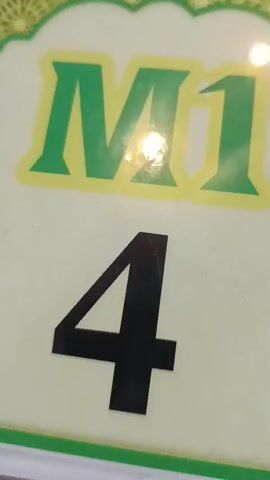 This is a close-up shot of a table marker. The marker is light cream-colored with a green border. At the top of the marker, there is a decorative green and yellow border with a repeating pattern. Below this, "M1" is printed in large, bold, green letters with a yellow outline. Beneath "M1", the number "4" is printed in large, bold, black numbers. The marker is slightly tilted, and there is a bright reflection on its surface. The background is blurred but appears to be a table with some other objects visible at the bottom edge.
