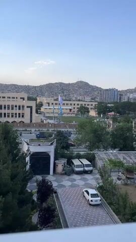 The video shows a view of a city skyline and surrounding hills from a balcony. In the foreground, a white car is parked in a courtyard with a patterned tile floor. To the left, there are two white vans parked next to a building with a large dark doorway. Behind these, a modern building with blue windows and the text "SCHOOL OF GRADUATE STUDIES" is visible. In the background, a densely populated hillside is covered with buildings, and a clear blue sky stretches above. The overall impression is of an urban environment with hilly terrain.