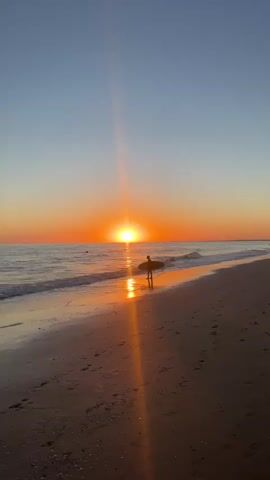 A lone person holding a surfboard walks along a sandy beach at sunset. The sun, a bright orange orb, is setting over the ocean, casting a shimmering reflection on the water. The sky is a gradient of orange and blue, with a single lens flare extending from the sun. Small waves lap at the shore, and footprints are visible in the sand. The scene is peaceful and serene.