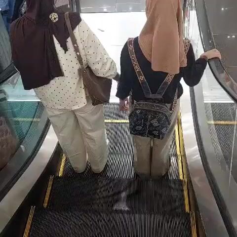 The video shows two individuals from the waist down, ascending an escalator. The first individual, on the left, is wearing a patterned white shirt, light-colored pants, and a brown headscarf. They are carrying a brown handbag over their left shoulder. The second individual, on the right, is wearing a black top, tan pants, and a beige headscarf. They have a dark patterned bag slung across their chest. Both individuals appear to be wearing dark shoes. The escalator steps are black with yellow accents. The background is slightly blurred, but hints of a retail environment can be seen, with polished floors and display shelves with what appear to be luggage in the upper right portion of the frame. The lighting suggests an indoor setting, likely a shopping mall or department store, during daylight hours. There is no visible text in the video. The mood is neutral and observational.