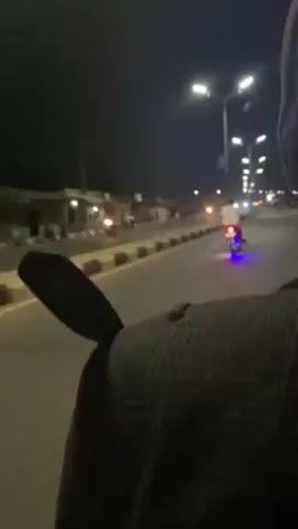 A person is recording video while riding a motorcycle at night. The road is asphalt with a concrete median, and is lit by tall streetlights. There are vehicles with headlights and taillights traveling on the road ahead, including at least one motorcycle with blue underglow lights. Small buildings line the side of the road to the left. The person recording is wearing a grey and black patterned jacket and a black knit hat.