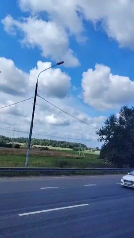 The video shows a view from a moving vehicle looking towards the side of a road. The road is paved and has white lane markings. To the right of the road, there is a grassy area and then a line of trees. Some of the trees are tall and leafy, while others are thinner and appear to be saplings. In the distance, there are fields and more trees, with a few power lines visible. The sky is blue with scattered white clouds. The overall impression is a rural or semi-rural landscape on a sunny day.