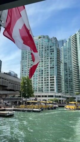 A Canadian flag is partially visible in the foreground, fluttering in the breeze. Beyond the flag, a body of water with rippling green water is seen. Several yellow boats, identified as "WATER TAXI," are docked or moving on the water. In the background, a row of modern, tall buildings with many windows stands against a bright blue sky with scattered clouds. The scene appears to be a waterfront area, possibly a harbor or marina, in a city.