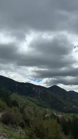 The video pans across a valley surrounded by mountains and a village nestled in the center. The sky is filled with thick, gray clouds, though patches of blue are visible. The mountains are covered in dense green forests. The village consists of closely packed buildings with red tile roofs.