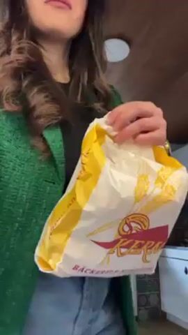 A young woman, from the waist up, wearing a black top, a green jacket with gold buttons, and blue jeans is shown in front of a light brown ceiling and a white cabinet. She opens a large white paper bag with yellow wheat designs on the sides and the brand name “Kern” in red. She takes out something green from the bag. She looks focused on her task.
